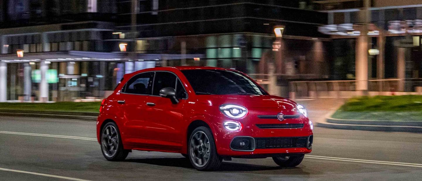 Three-quarter side view of a red 2020 Fiat 500X being driven on a downtown street at night.